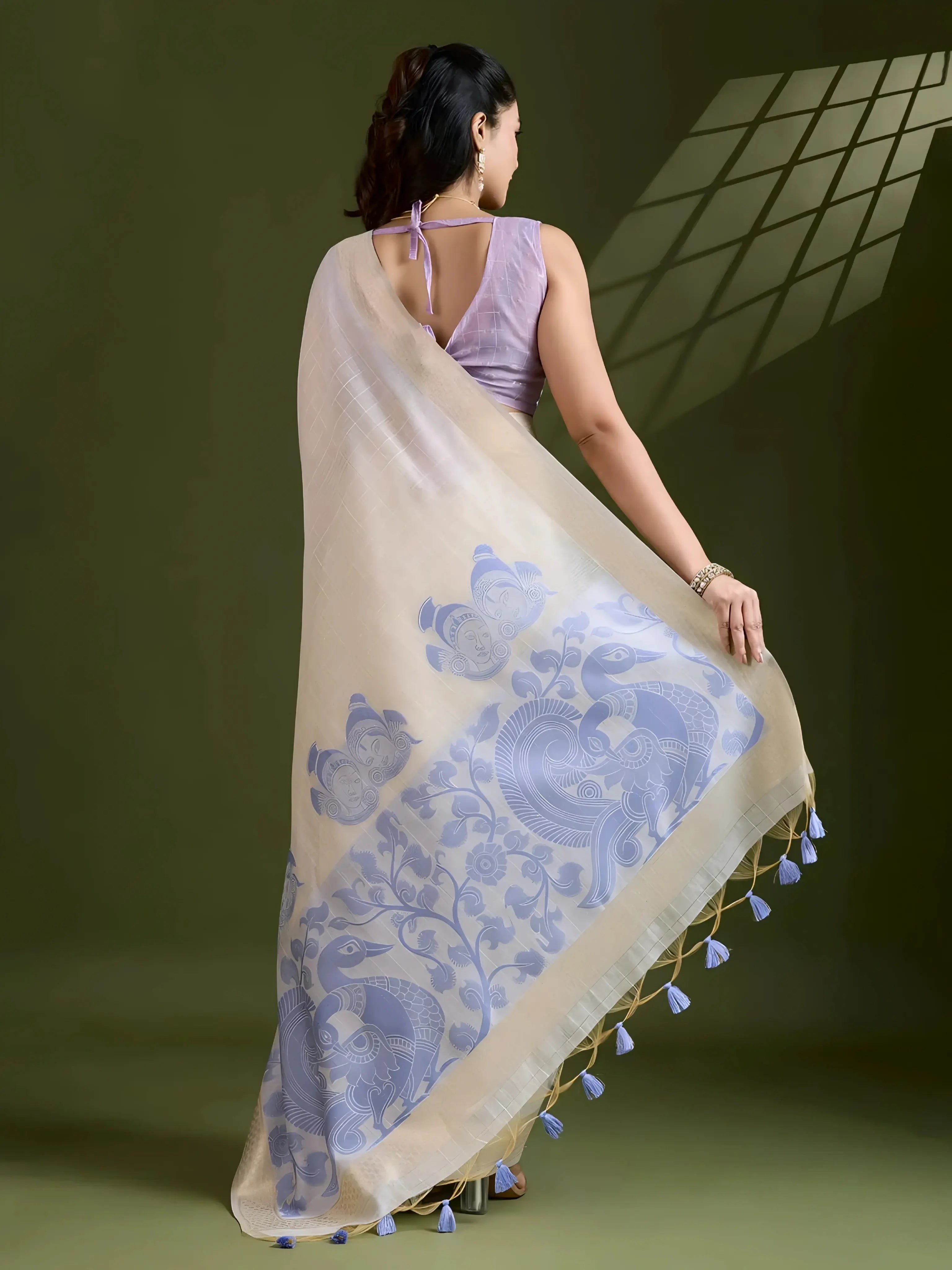 A woman models an elegant Beige Soft Muga Cotton Saree. The saree features large Kalamkari-style prints of peacocks and florals in a contrasting Soft Lavender Blue on the pallu. She wears a sleeveless lavender blouse and accessorizes with a gold and pearl Kundan choker necklace, posing indoors.